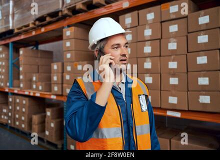 Male technician talking on smartphone wearing hardhat at manufacturing storehouse Stock Photo