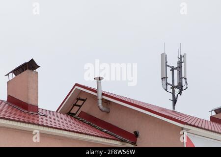 Telecommunication antenna on the roof, cellular and telephone communications. Smoke and ventilation pipes against the gray sky. Red tiles on the roof of a residential building. Stock Photo