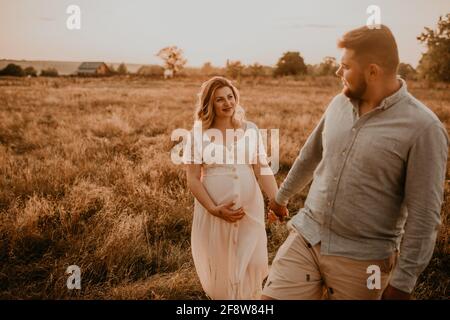 Happy family pregnant caucasian blonde woman with moles on face in white cotton dress walks with husband meadow summer. man in light natural clothes a Stock Photo