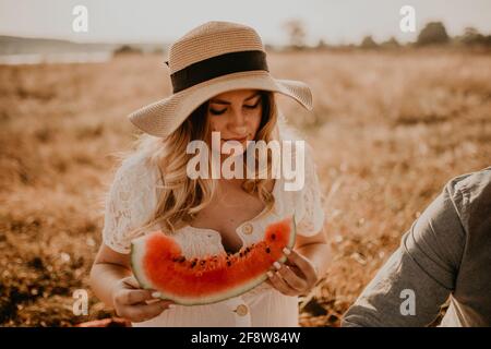 happy European caucasian pregnant woman relaxing in nature picnic eating fruit red juicy watermelon laugh having fun. expectant mother in hat and dres Stock Photo