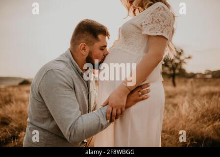 husband hugs pregnant wife hold hands on big round tummy kissing belly. Happy family resting in nature hugs kisses in summer at sunset. future mother Stock Photo