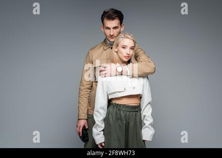trendy man holding binoculars and hugging blonde woman isolated on grey Stock Photo