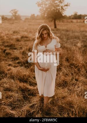 Caucasian pregnant young blonde woman in cotton white linen dress stand walking meadow on dry grass in summer at sunset nature. mother-to-be holds her Stock Photo