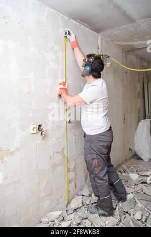 Builder man using a measuring tape at the construction site. Construction worker wearing safety gloves, glasses and earmuffs. Stock Photo