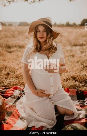 happy European caucasian pregnant woman relaxing in nature picnic looking at camera. expectant mother in hat and dress hold hand on belly. face with m Stock Photo