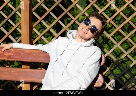 Portrait of pretty young adult caucasian woman in white hoodie sweater and sunglasses enjoy sunbathing warm sunlight at yard home garden outdoors Stock Photo