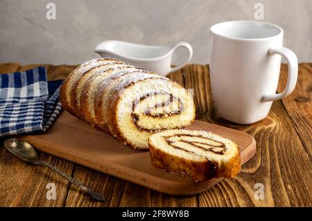 Uncut biscuit roll with fruit jam and powdered sugar on a cutting board, with a white tea cup. Homemade baking. Breakfast. Stock Photo
