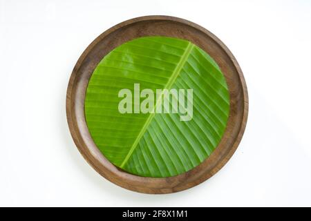 Banana leaf,fresh green banana leaf  cut in circular shape and placed on a wooden plate with white  textured background ,isolated. Stock Photo