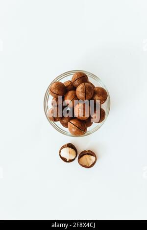 Raw macadamia nuts in a transparent glass bowl and scattered on grey solid background. Minimal composition. Stock Photo