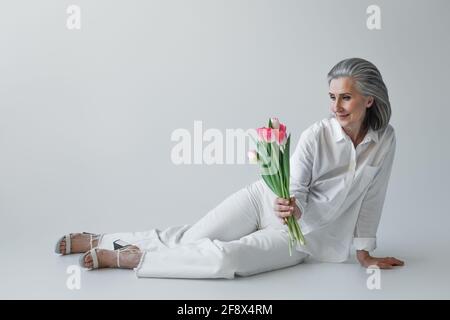 Smiling woman in white clothes holding tulips on grey background Stock Photo