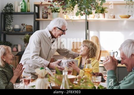 Senior man giving present to senior woman while they sitting at the table and having dinner at home Stock Photo