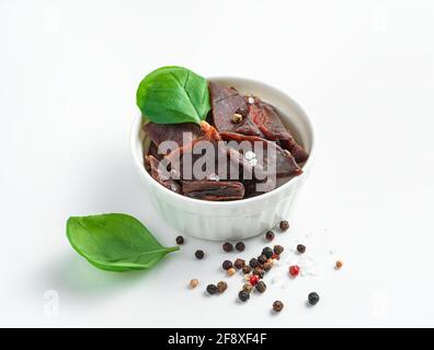 Savory appetizer, dried meat with pepper and salt in a white cup decorated with basil on a white background. The concept of natural snacks. Stock Photo