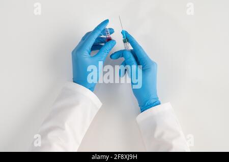 A nurse on a white background in sterile gloves holds a vial with serum and a syringe for injecting sick patients. Stock Photo