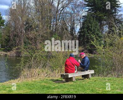 An elderly couple sitting on a bench by the River Thames enjoying a snack together on a sunny spring day Runnymede Surrey England UK Stock Photo