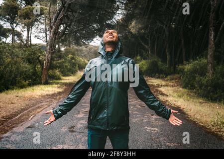 Young wet smiling man in drenched jacket with a hood enjoying rain. Man in raincoat stood in heavy rain. Bad autumn wether. Stock Photo