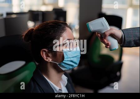 Business woman in medical mask has body temperature measured with electronic thermometer at workplace in office. Stock Photo