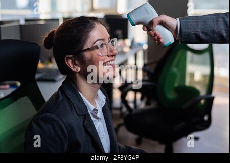 Smiling business woman has body temperature measured with electronic thermometer at workplace in office. Stock Photo