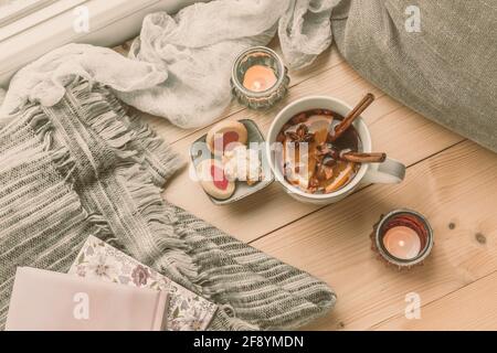 Tea and biscuits on a wooden floor in front of a window with pillows and a woolen blanket, concept for cozy winter afternoon at home Stock Photo
