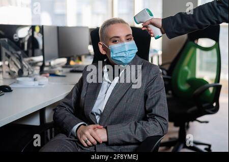 Business woman in medical mask has body temperature measured with electronic thermometer at workplace in office. Stock Photo