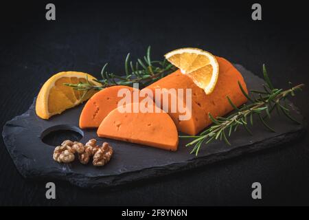Semicircular piece of orange cheese on a slate, garnished with orange, walnuts and rosemary Stock Photo
