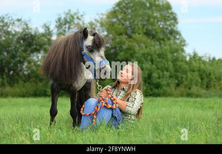 Young Caucasian woman is sitting on the grass next to her small horse are in outdoors. Stock Photo