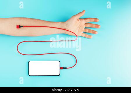 The hand is connected by a red power cord to a smartphone with free space on a blue background. The concept of drug addiction on the social networks o Stock Photo