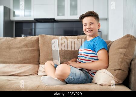 Smiling teenager in casual outfit sitting with crossed legs on couch and looking at camera. Happy boy using modern laptop while staying at home. Stock Photo