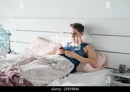 Young man in female shirt lying on bed and using mobile phone in the morning in his bedroom Stock Photo