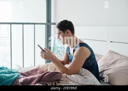 Young man in female clothes sitting in his bed and texting a message to his boyfriend in the morning at home Stock Photo