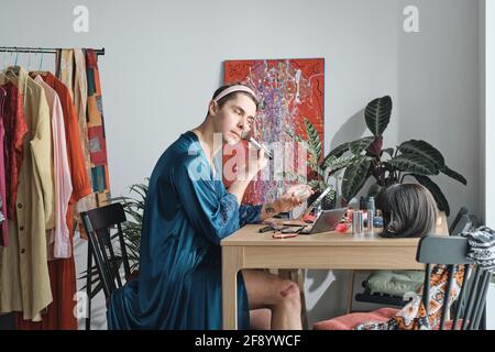 Young transsexual sitting at the table and applying makeup on his face in dressing room Stock Photo