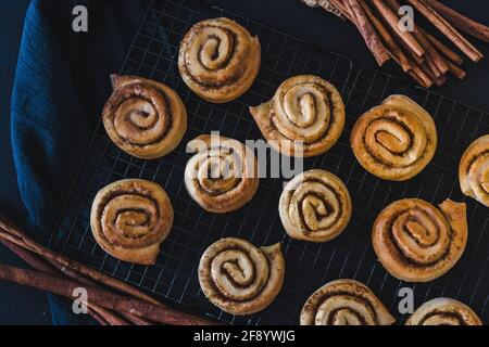 Homemade cinnamon rolls on a cooling rack with cinnamon sticks on dark blue background. Stock Photo