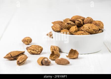 Roasted and salted snack almonds with shell in a small white bowl on white wooden background, copy space Stock Photo