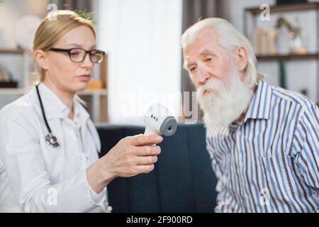 Digital infrared thermometer in hand of pleasant blond 40-aged woman doctor or nurse, visiting her senior male patient at home, showing him the results of body temperature measurement. Stock Photo