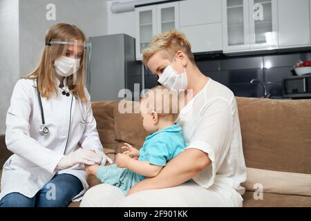 Professional doctor in medical uniform and mask checking health condition of little baby. Young mother holding boy on knees during medical examination. Stock Photo