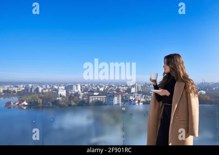 Side view of young beautiful brunette woman in beige coat drinking coffee or tea and looking through panoramic window. Concept of free time with hot drink. Stock Photo