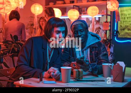 Young man sitting at the table with his boyfriend they watching something on mobile phone sitting in nightclub Stock Photo