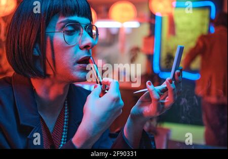Young man in short black wig applying lipstick and looking at mirror while sitting in the nightclub Stock Photo