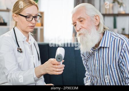 Close up portrait of pleasant blond 40-aged woman doctor or nurse, visiting her senior male patient at home, showing him the results of body temperature measurement on digital infrared thermometer Stock Photo