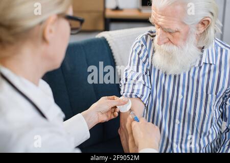 View from the shoulder of female professional doctor. Senior bearded man, is vaccinated at home by his family gp doctor. Female doctor makes an insulin injection to male diabetic patient at home. Stock Photo