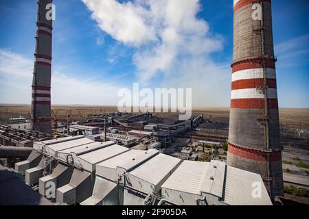 GRES-1 thermal power station. grey raw coal bunkers on front. Smoke stacks both sides.Coal-harvesters loading coal to electricity plant.Dry grass and Stock Photo