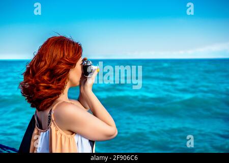 Red haired woman in summer dress travel photographer, taking pictures of sea. Rare photographic camera for vacation snapshots. Stock Photo