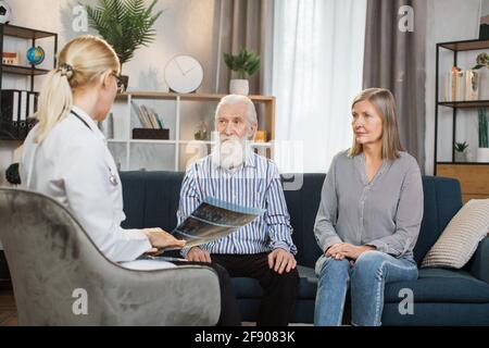 Retired couple sitting on the sofa at home and listening to their gp doctor. Young high-skilled female healthcare worker, holding CT x-ray scan, consulting joyful senior couple during home visit. Stock Photo
