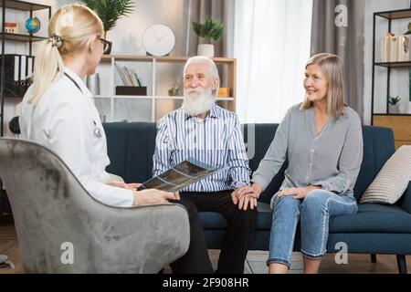 Young high-skilled female healthcare worker, holding CT x-ray scan, consulting joyful senior couple during home visit. Retired couple sitting on the sofa and listening to the doctor. Stock Photo