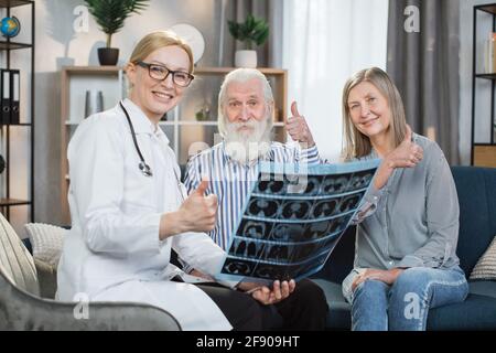 Good news, strong health. Pretty smiling female doctor satisfied of the results of x-ray scan of her patients, happy bearded senior man and pleasant woman , showing thumbs up to camera. Stock Photo