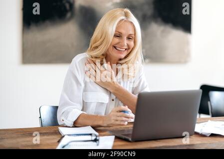 Friendly, confident elegant mature blonde, business lady or manager, uses laptop, chatting on video conference with coworkers or client, gesturing with hand and smiling. Online video meeting Stock Photo