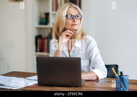 Attractive successful caucasian senior business woman, blond lady, wearing a white shirt and glasses, sits in front of laptop, taking break, looking pensively to the side, dreaming about vacation Stock Photo