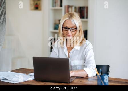 Concentrated influential beautiful caucasian blonde mature woman, wearing glasses and white shirt, ceo, business woman or broker working on laptop, making report, develop strategy, typing on keyboard Stock Photo