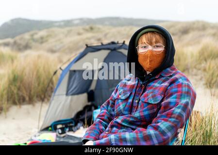 Teen in camp chair on the coast with mask looking at camera near dunes Stock Photo