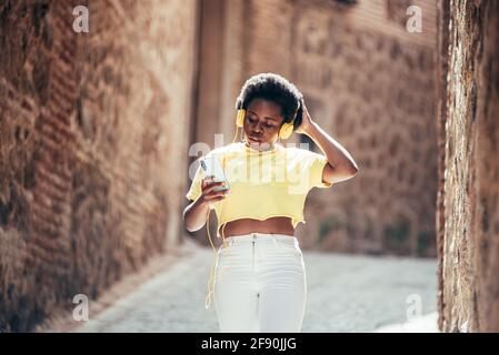 Portrait of black girl with afro hair listening to music with headphones and her mobile phone while walking down an old city street. Stock Photo