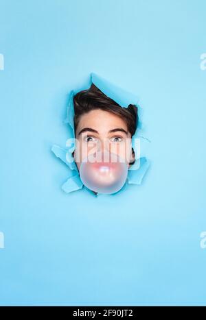 Boy blowing bubble with head through hole in blue paper background. Stock Photo